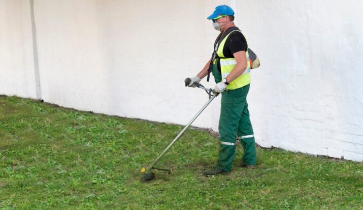 Mężczyzna w kamizelce odblaskowej i maseczce ochronnej koszący trawę przy ścianie budynku za pomocą kosy spalinowej.